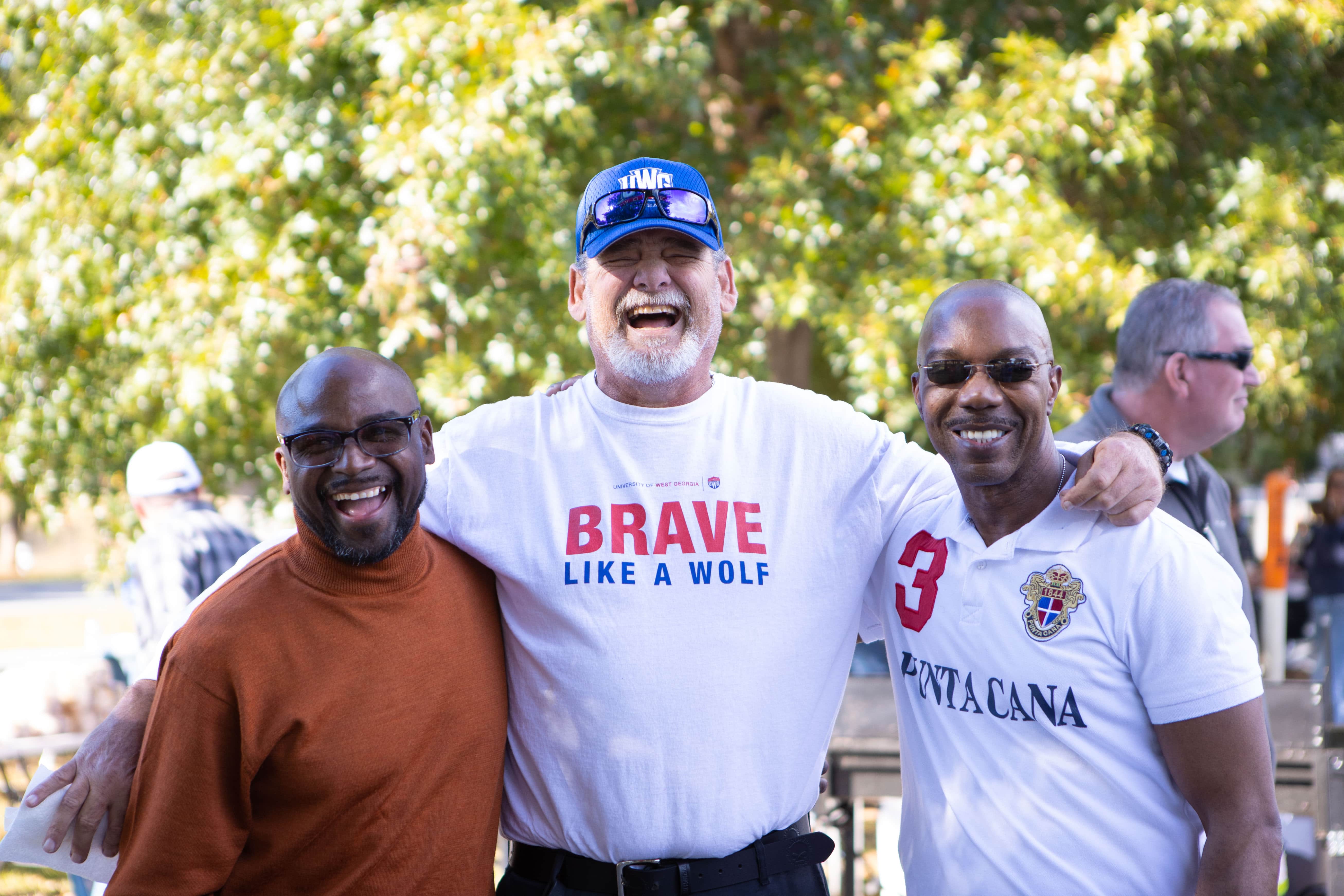 Three 沙巴官网 alumns standing together for a photo. The center person is wearing a white shirt with "Brave Like a Wolf" written on the front, and a 沙巴官网 hat.